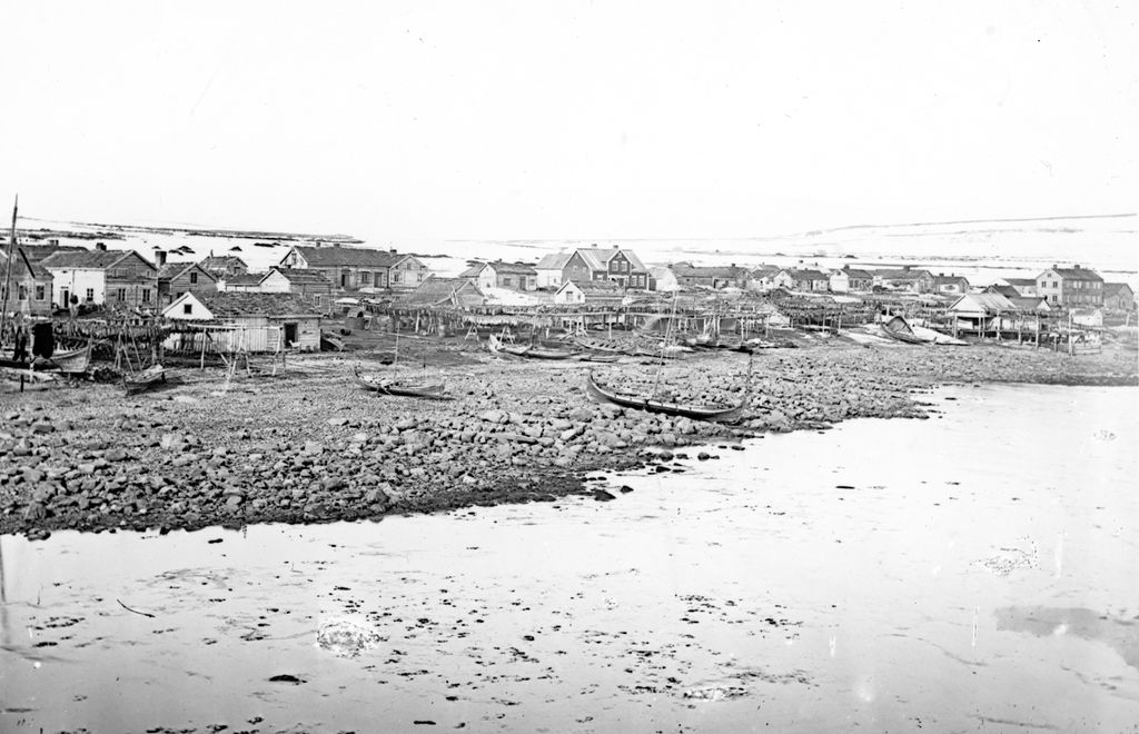 Vadsø på slutten av 1800-tallet. Trehusbebyggelse ved sjøen. Noen få båter i forgrunnen. Foto.