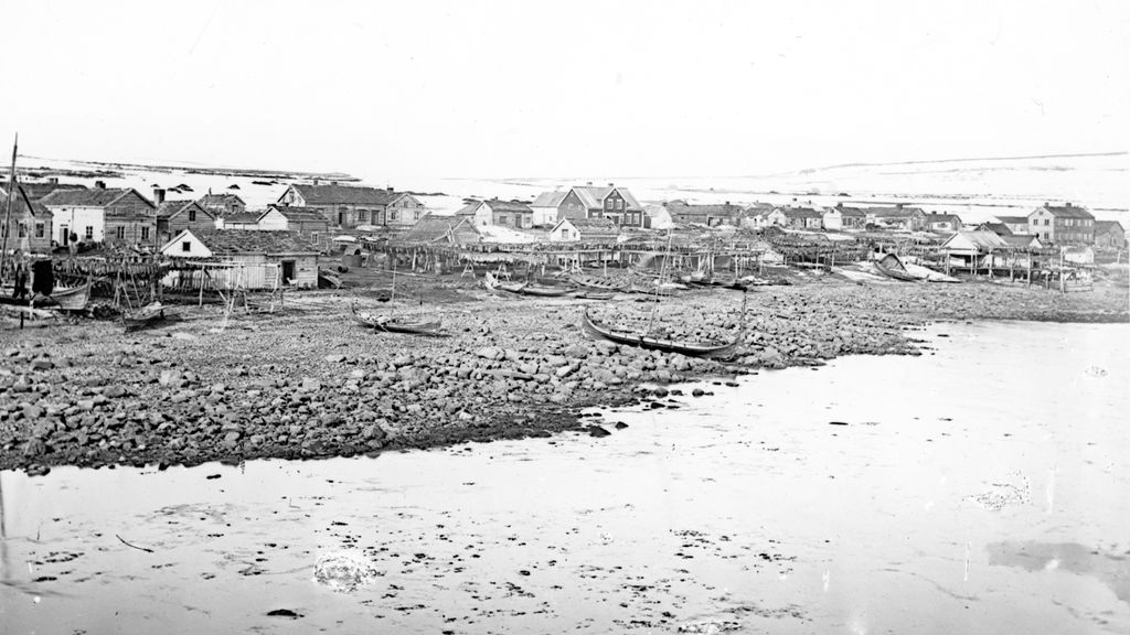 Bebyggelse ved sjøen. Bebyggelsen er preget av trehus, noen med en etasje og noen med to etasjer. Svart-hvitt foto.