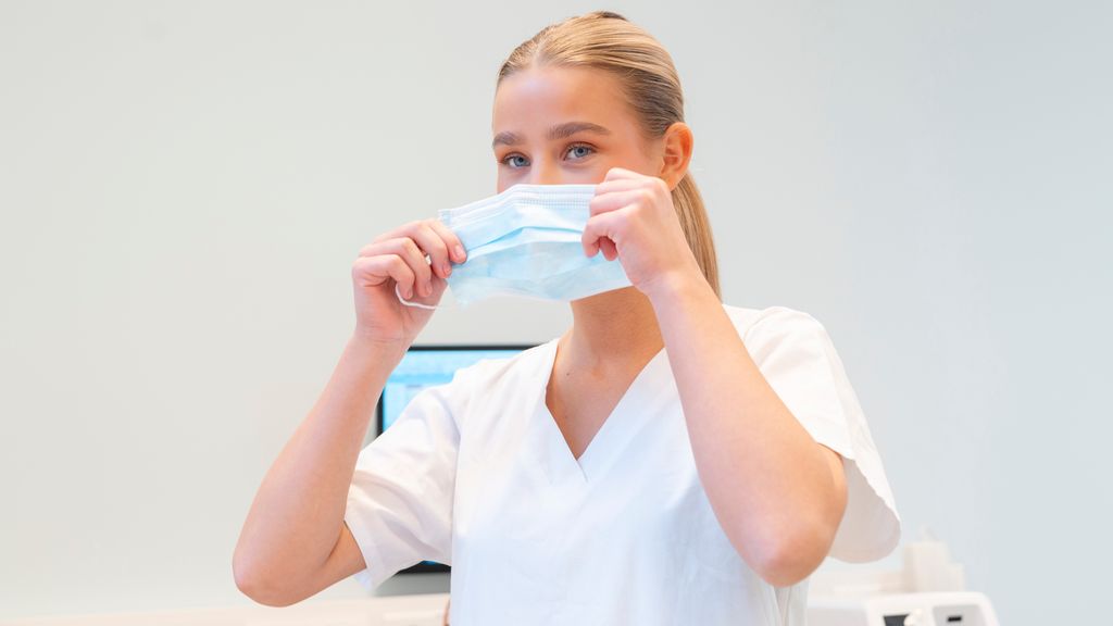 Kvinne i uniform for helsepersonell tar på munnbind. Foto.