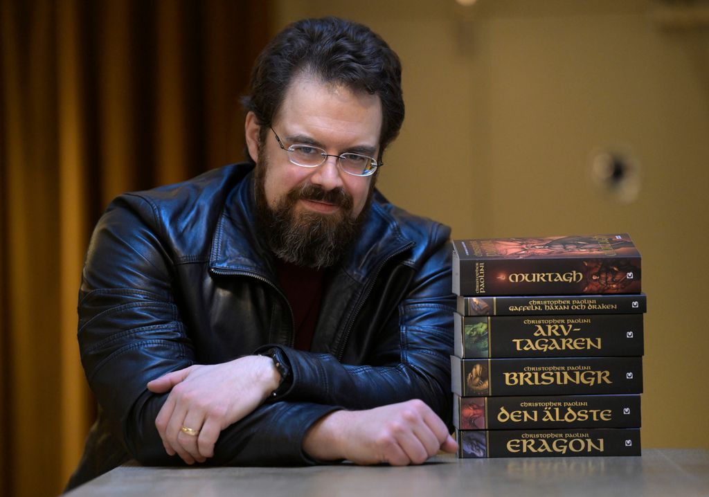 Fantasyforfatteren Christopher Paolini sitter ved et bord. Det er en stabel med bøker på bordet. Foto.