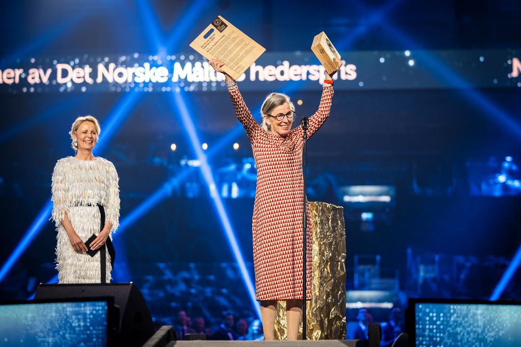 Jublende kvinne på en scene løfter utmerkelser i været. Bak henne står en annen kvinne. Bak dem sitter publikum, og på et lysbanner står det Det Norske Måltids hederspris. Foto.