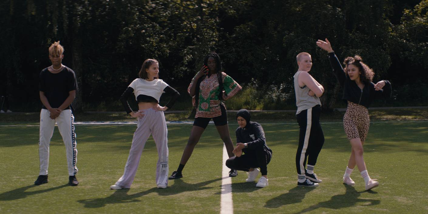 Utsnitt frå filmen "Alle utlendinger har lukka gardiner". Fem jenter i ulike typar fritidsklede står på ei kunstgrasbane. 