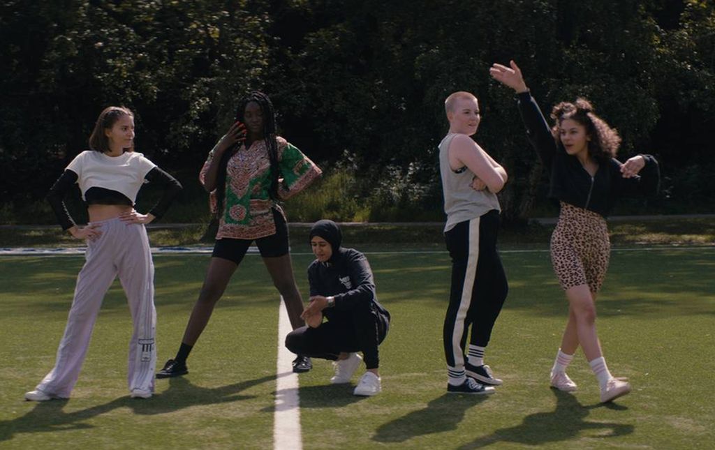 Utsnitt frå filmen "Alle utlendinger har lukka gardiner". Fem jenter i ulike typar fritidsklede står på ei kunstgrasbane.