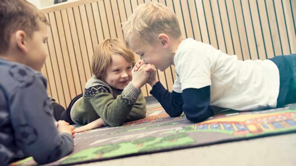 To gutter ligger på et bilteppe på gulvet og bryter håndbak. En tredje gutt ligger ved siden av og ser på. Foto.