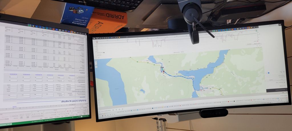 Et svart skrivebord med to store dataskjermer. I forgrunnen et tastatur og hodetelefoner. I bakgrunnen ADR-handboka og en lastsikringsbrosjyre. Foto.