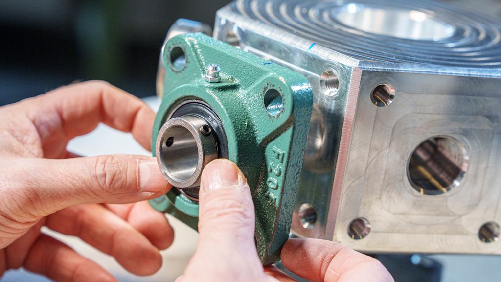 To hender holder en grønn, firkanta metallkonstruksjon mot ei sølvfarga festeplate. Foto.