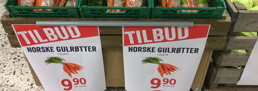 Ei grønnsakshylle i en butikk. På hylla henger det plakater med informasjon om tilbudspris på norske gulrøtter. Foto.