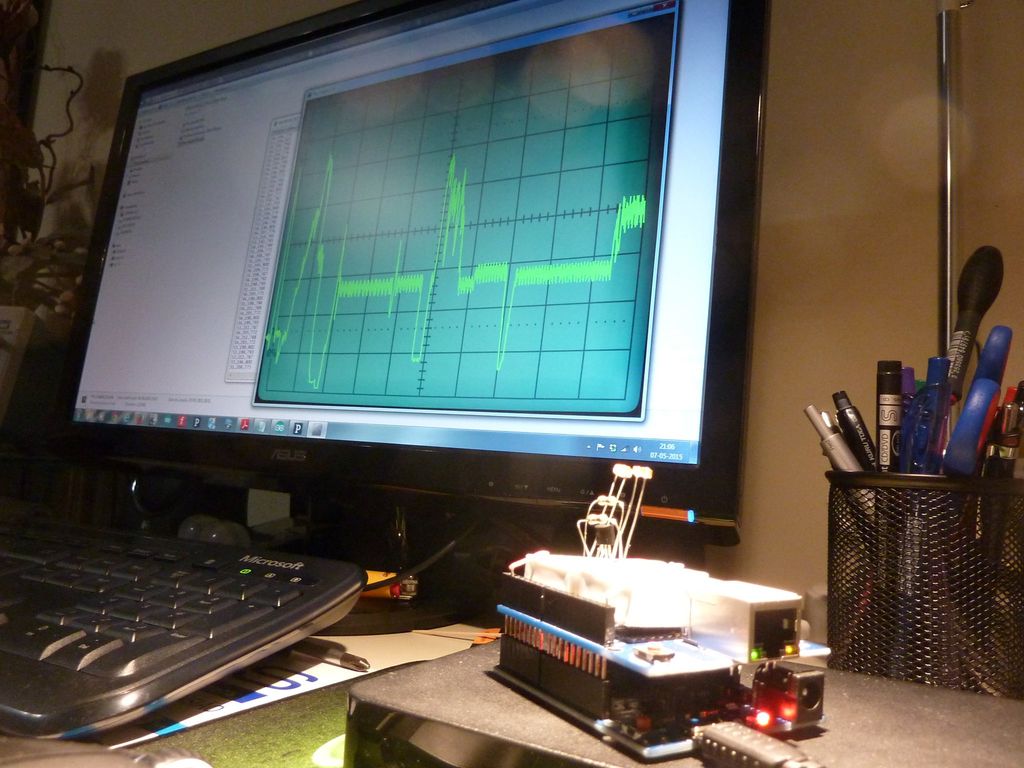 En dataskjerm med tastatur der skjermen viser grafisk framstilling av oscilloskopsignaler. En elektronisk enhet er koplet til en pc. Foto.