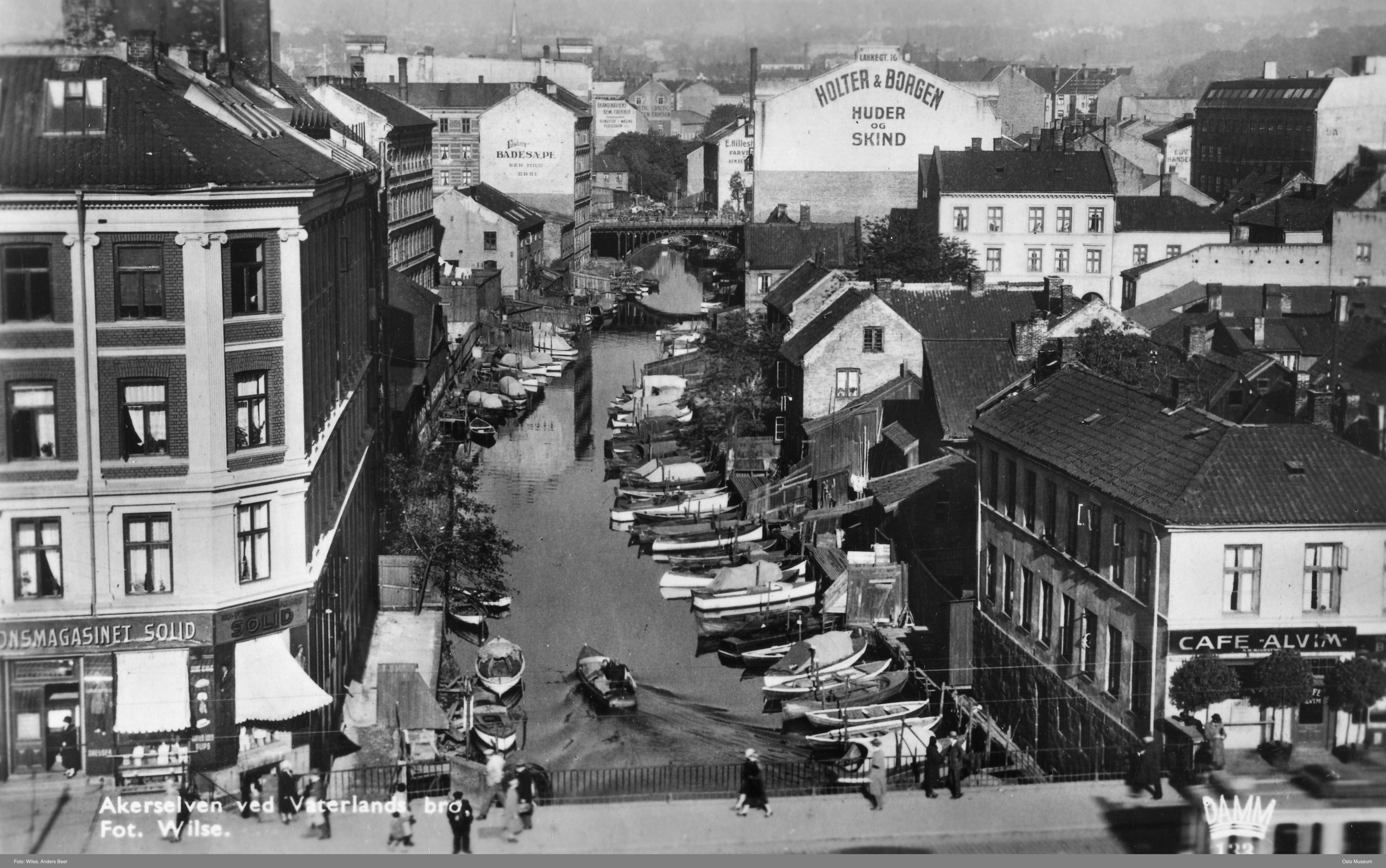 Akerselva ved Vaterlands bru der elva flyt sakte gjennom byen. Små båtar ligg fortøydde tett i tett  langs elva. Foto.