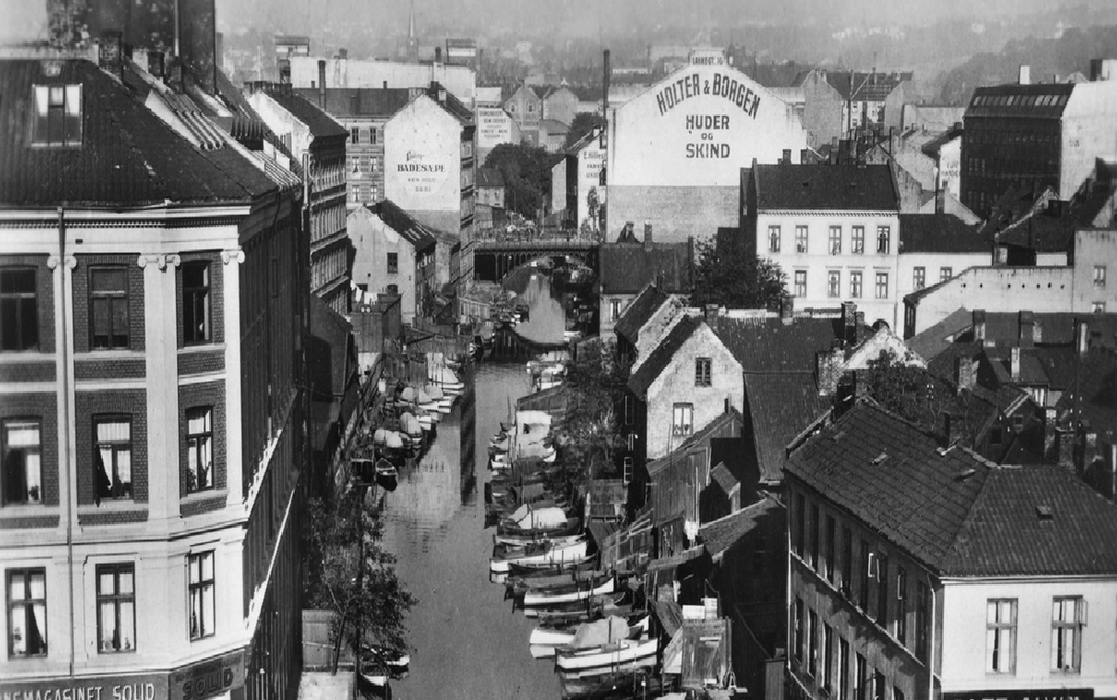 Akerselva ved Vaterlands bro hvor elva flyter sakte gjennom byen. Små båter ligger fortøyd tett i tett langs elva. Foto.
