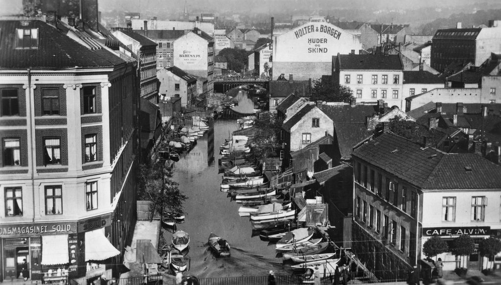 Akerselva ved Vaterlands bro hvor elva flyter sakte gjennom byen. Små båter ligger fortøyd tett i tett langs elva. Foto.