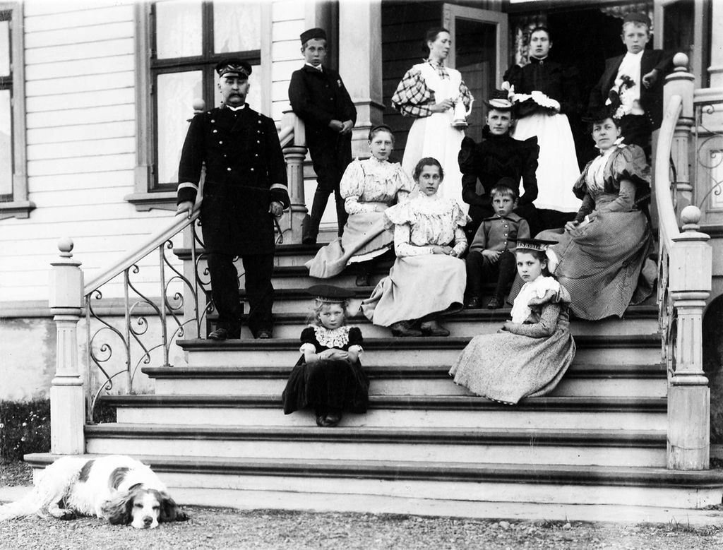 En familie står og sitter på trappen til et hus. Familien er kledd i finere klær. Noen på bildet er sannsynligvis hushjelper. Det ligger en hund nedenfor trappen. Huset ser stort ut, og trappen har flere trinn med forseggjort rekkverk. Bildet er fra Vardø i 1898 og viser amtmann Graff med familie og hushjelper. Svart-hvitt-foto.