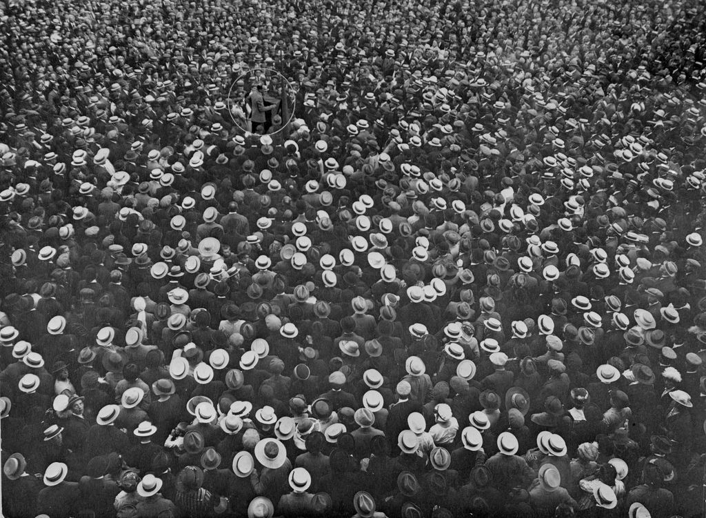 Et utendørsarrangement der en stor folkemasse står tett i tett rundt en mannlig taler ved et podium. Flertallet i forsamlinga er menn. Alle, også kvinnene, bærer hatter. Foto. 