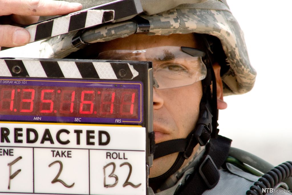 Mann med hjelm bak en filmklapper. Foto.