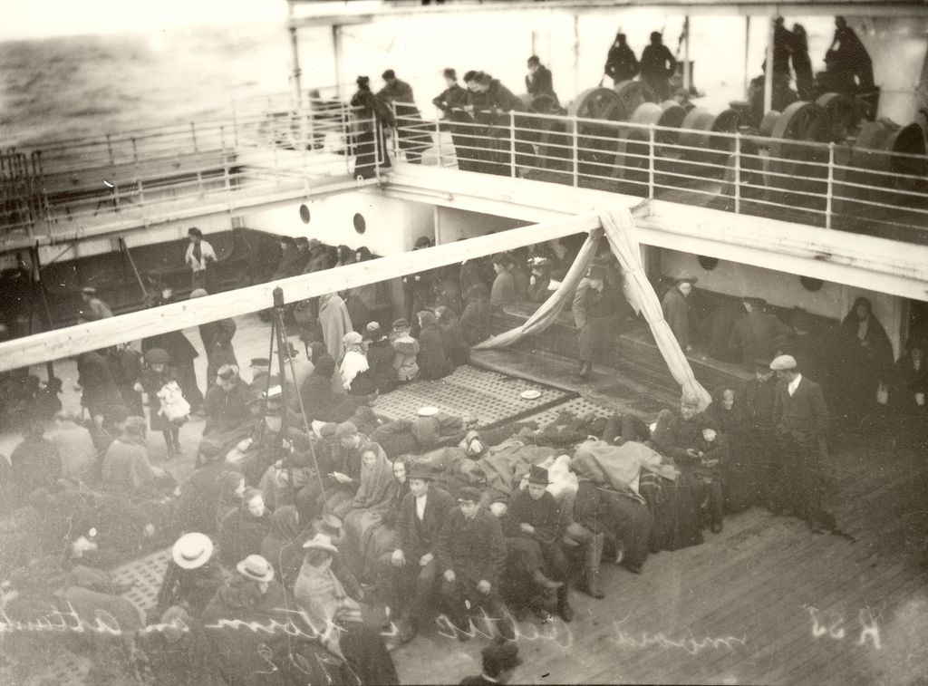 Mange passasjerer ute under åpen himmel på et skip. Noen står og andre sitter. Foto.