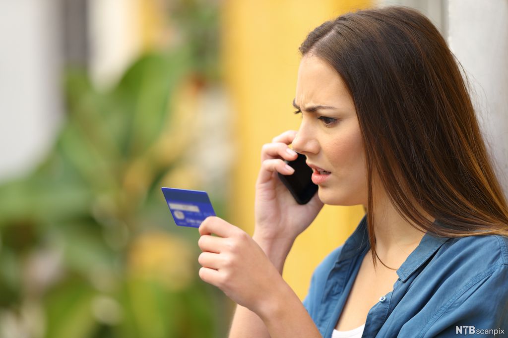 En sint dame holder et kredittkort i den ene hånden og en telefon i den andre. Foto.