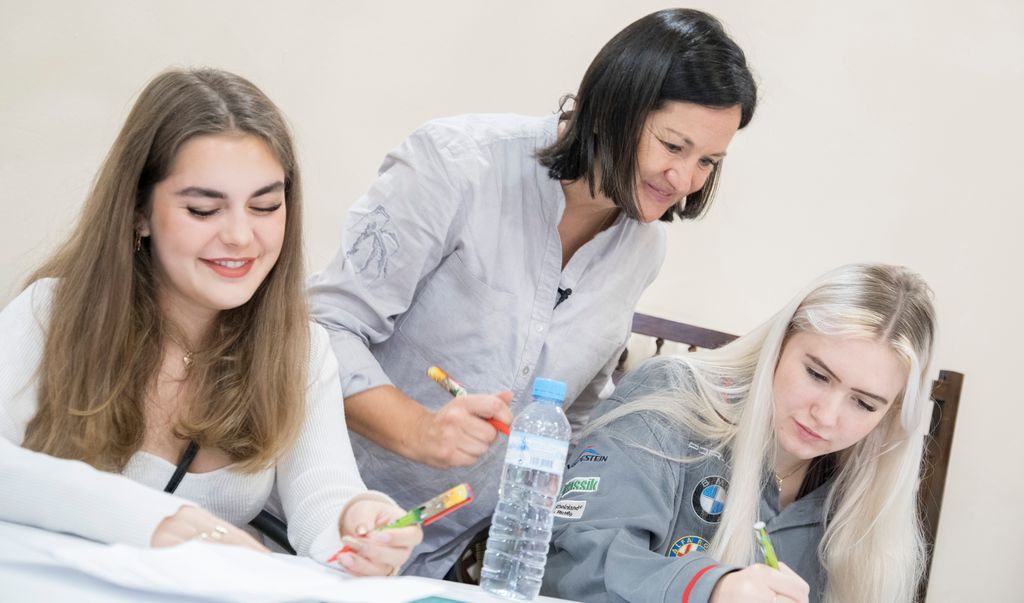 En lærer står bak to elever som sitter og jobber. Læreren ser på det elevene har skrevet. Foto.