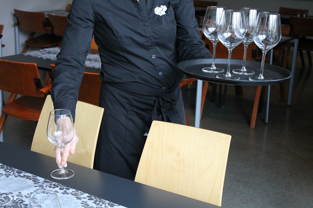 Servitør viser teknikk med å setje på plass stettglas. Hen held eit brett med glas i venstre hand og løftar eit glas ved å halde i stetten med høgre hand. Foto