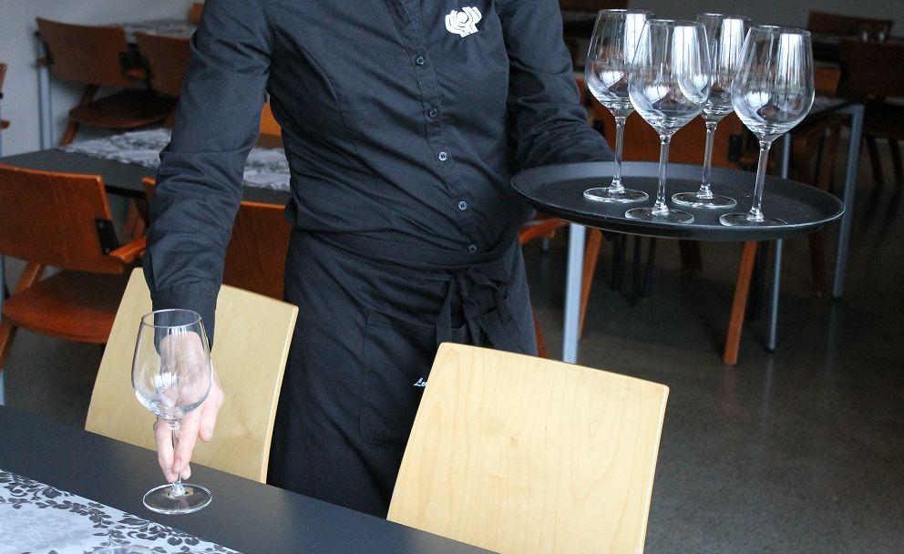 Servitør viser teknikk med å setje på plass stettglas. Hen held eit brett med glas i venstre hand og løftar eit glas ved å halde i stetten med høgre hand. Foto