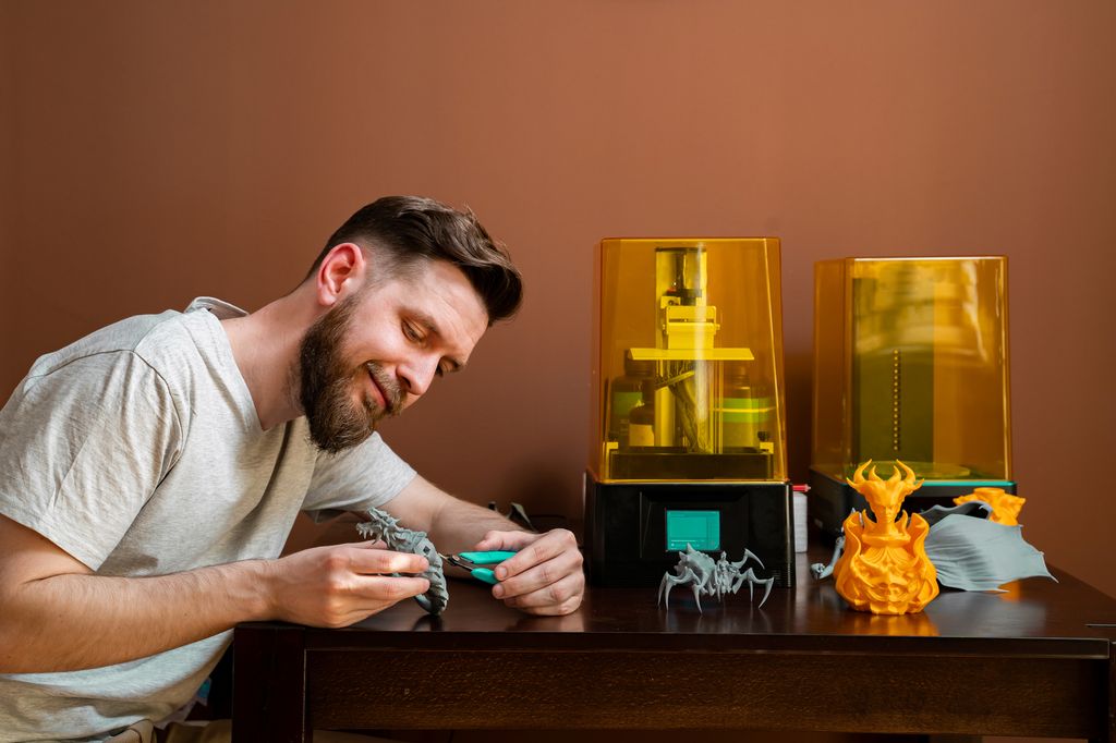 En person sitter ved et bord med to resinbaserte 3D-printere og flere ferdigprintede modeller. Personen holder en liten 3D-printet figur og utfører etterbehandling ved hjelp av håndverktøy. På bordet står ulike 3D-printede objekter med detaljerte overflater, klare for rengjøring, sliping eller videre bearbeiding etter printing. Foto.