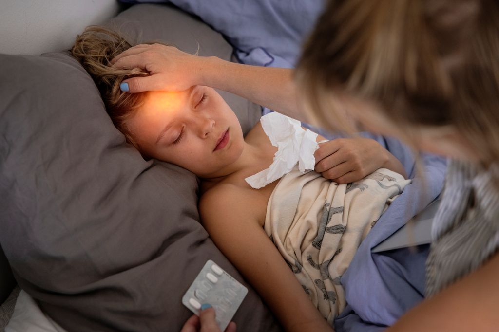 Et barn ligger i ei seng med lukkede øyne og holder på en papirserviett. En person holder ei hånd på panna til barnet, mens hun holder et brett med piller i den andre hånda. Foto.