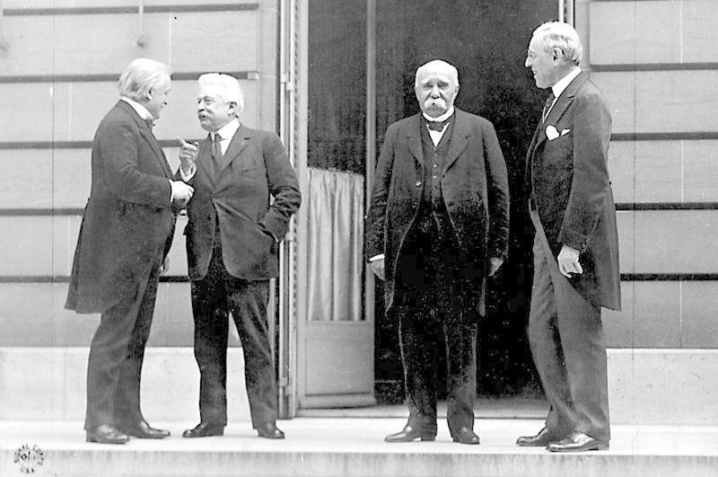 Fire menn med grått hår, kledd i mørke dresser. De står på en trapp med ei åpen dør i bakgrunnen. Foto.