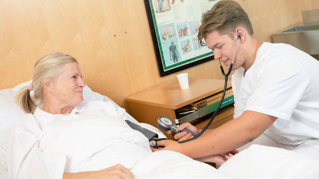 Mannlig helsefagarbeider måler blodtrykk på en sengeliggende kvinnelig pasient. Begge smiler. Foto.