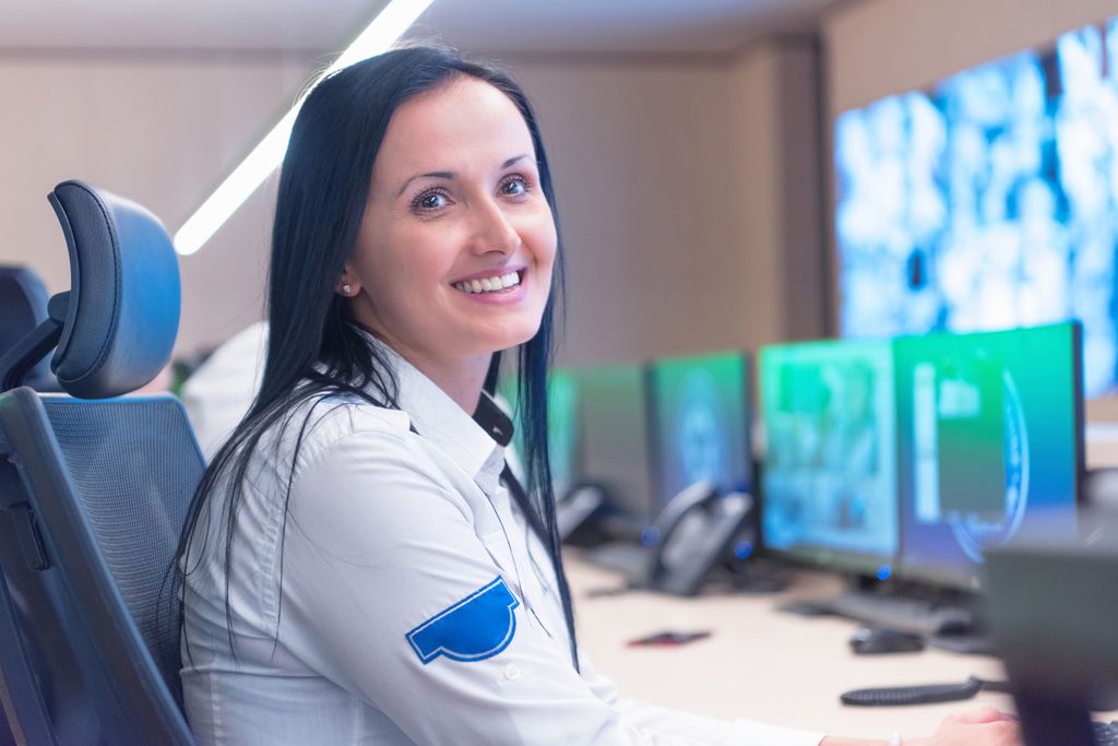 Uniformert kvinnelig vekter sitter ved en arbeidspult med overvåkingsskjermer og ser smilende inn i kameraet. Foto.