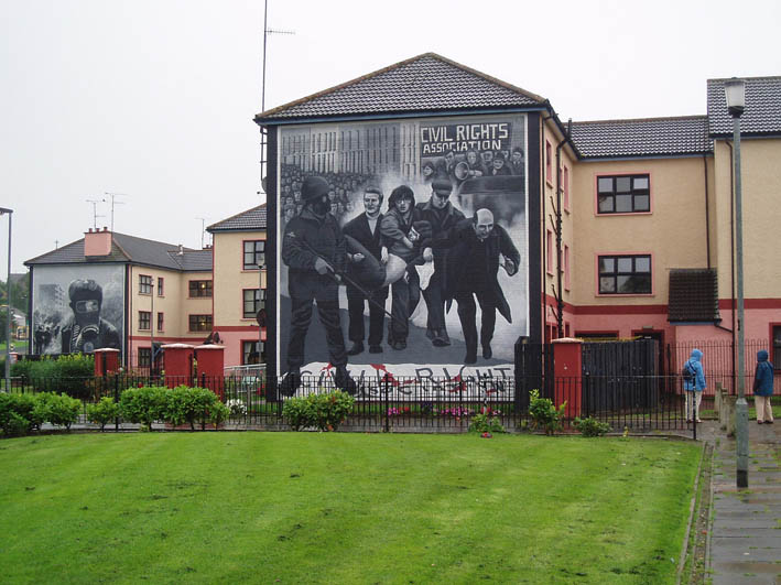 Photo: Mural commemorating Bloody Sunday. We see the words 'Civil Rights Association'. A soldier. People carrying a bleeding victim. Protesters in the background. 