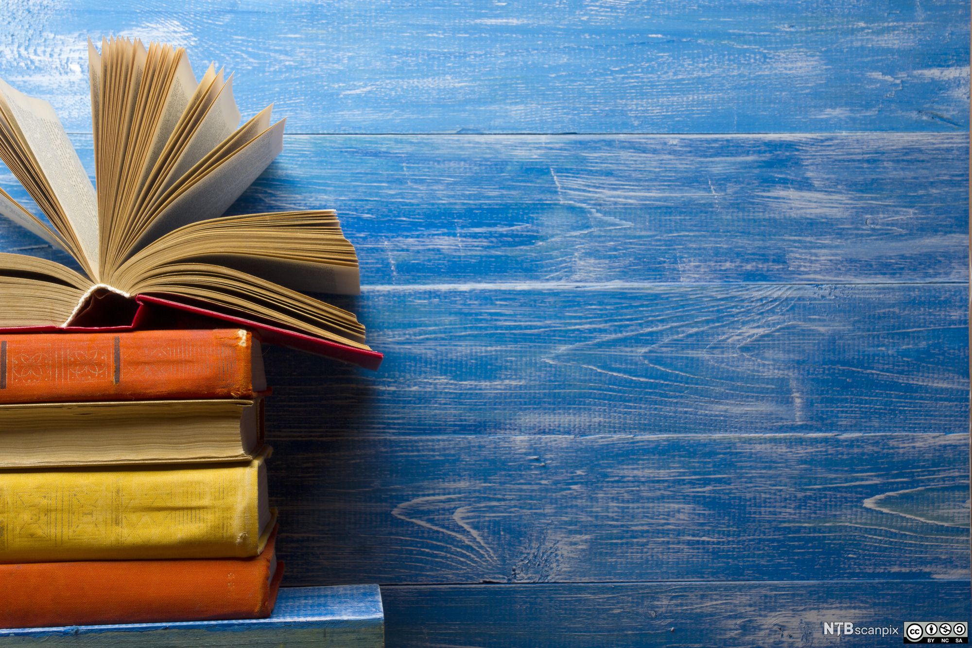 En stabel bøker hvor den øverste er åpna, foran en blå trevegg. Foto.