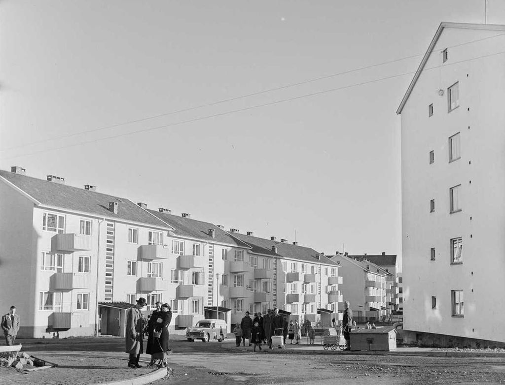 Mennesker ute mellom de tre etasjer høye boligblokkene. Mennene har hatt og frakk, et par barnevogner og én enkelt bil er også med på bildet. Svart-hvitt foto.