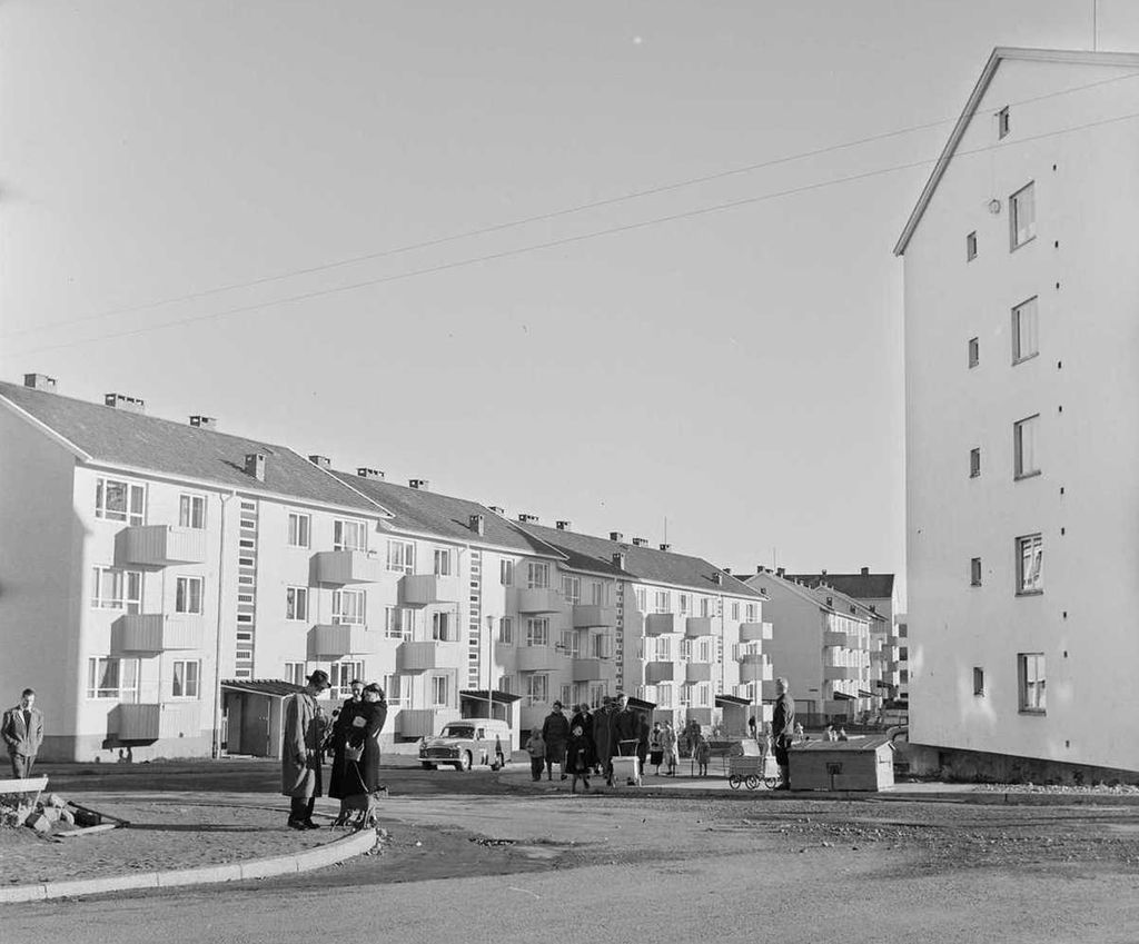 Mennesker ute mellom boligblokkene i drabantbyen. Det er treetasjes murhus med saltak. Til hver leilighet er det små balkonger. En enslig bil er med på bildet. Foto.