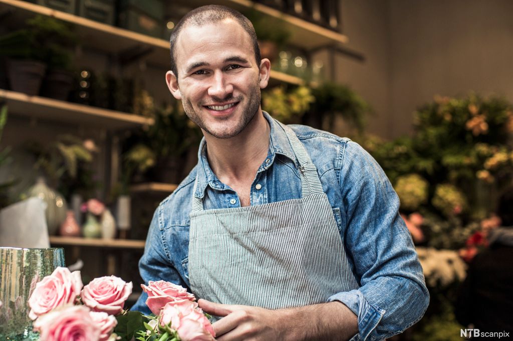Smilende mann i forkle omgitt av blomster. Foto.