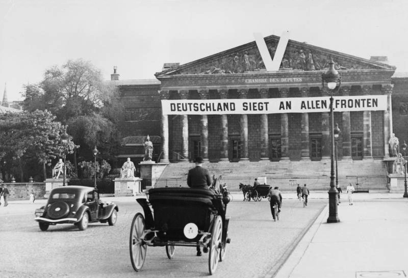 Nasjonalforsamlingen i Paris er forsynt med et tysk banner der det står "Deutschland siegt an allen Fronten". På gata foran er det en bil, to hester med vogn, et par syklister og noen gående. Foto. 