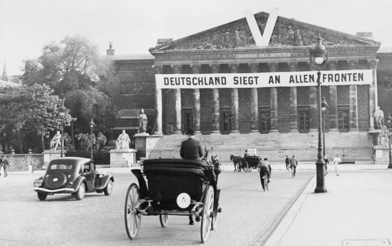 Nasjonalforsamlingen i Paris er forsynt med et tysk banner der det står "Deutschland siegt an allen Fronten". På gata foran er det en bil, to hester med vogn, et par syklister og noen gående. Foto.
