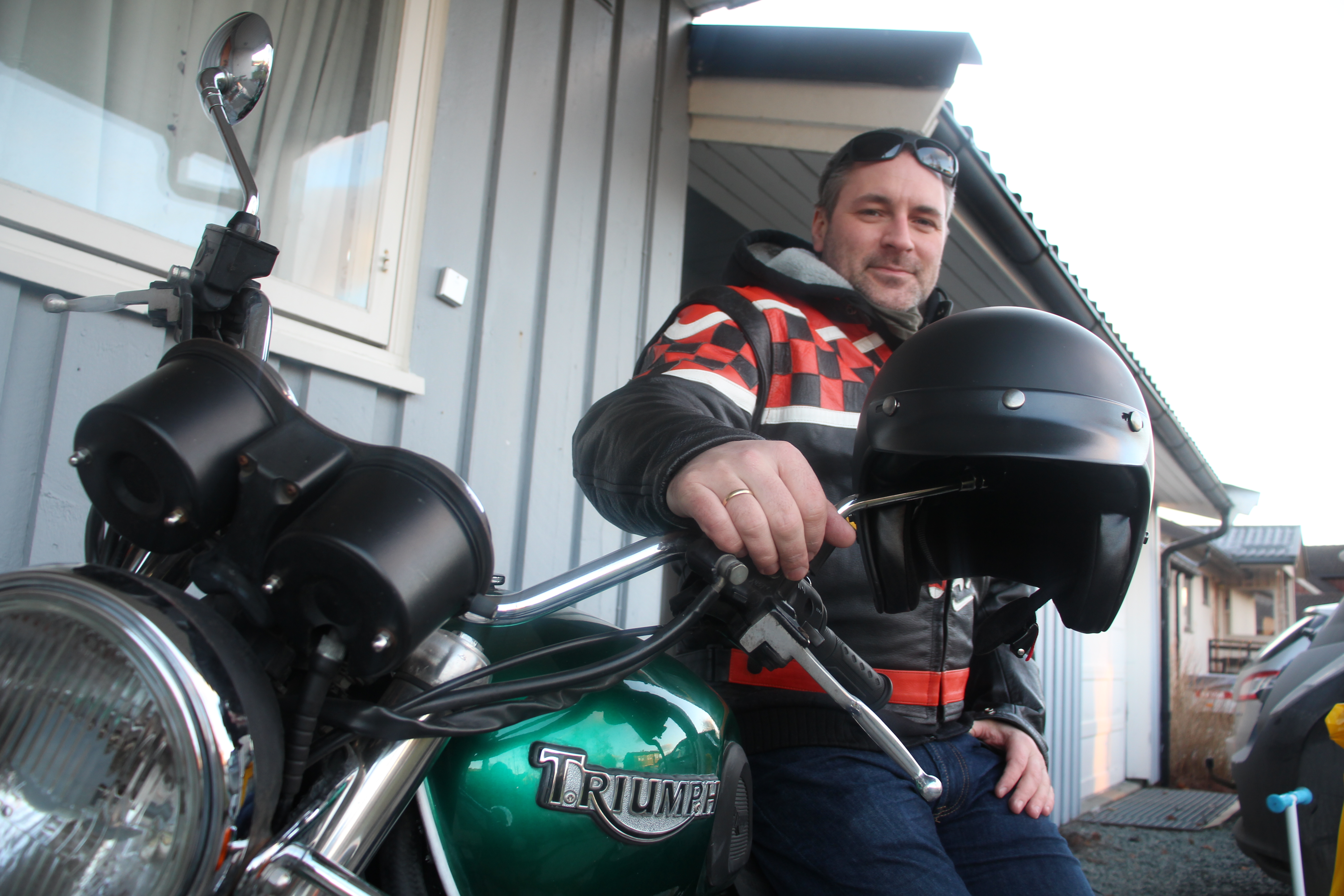 En mann med solbriller og motorsykkeljakke står ved den grønne motorsykkelen sin og ser mot kameraet. En svart hjelm henger på styret. Foto.