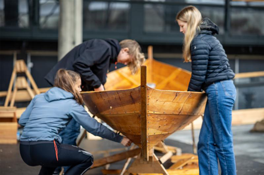 Tre elever arbeider på en liten trebåt som står inne i en hall. Foto. 