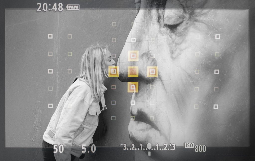 Et gatemaleri viser et nærportrett av en eldre person. Ei jente kysser personen på bildet på nesa. Fokuspunktene i kameraet er plassert på jentas munn og nesa til den gamle. Foto.