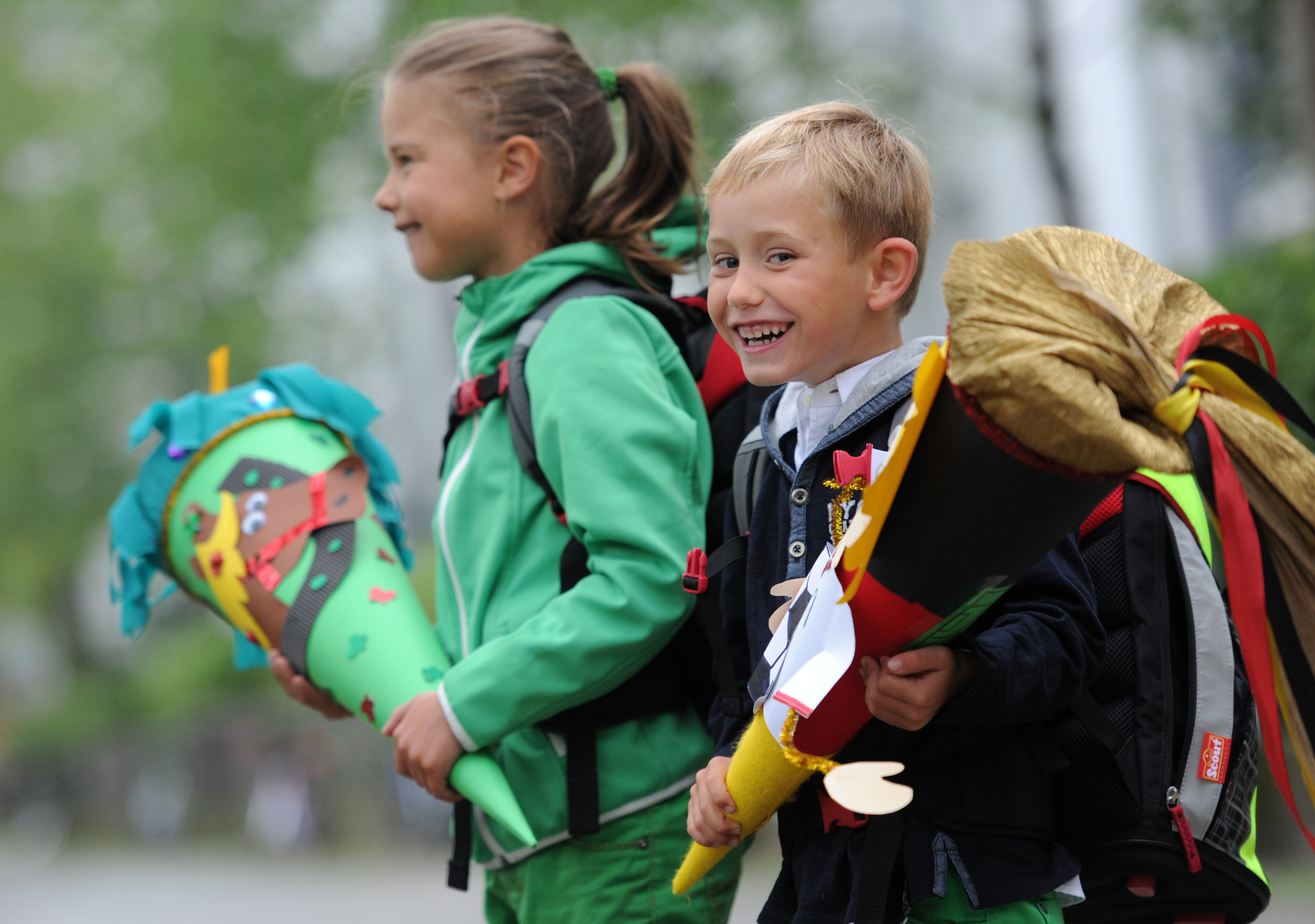 To smilande førsteklassingar, ein gut og ei jente, på veg til skulen første skuledag. Begge har ransel på ryggen og ber på enorme kremmerhus pynta med teikningar og pålimde motiv i mange fargar. Foto.