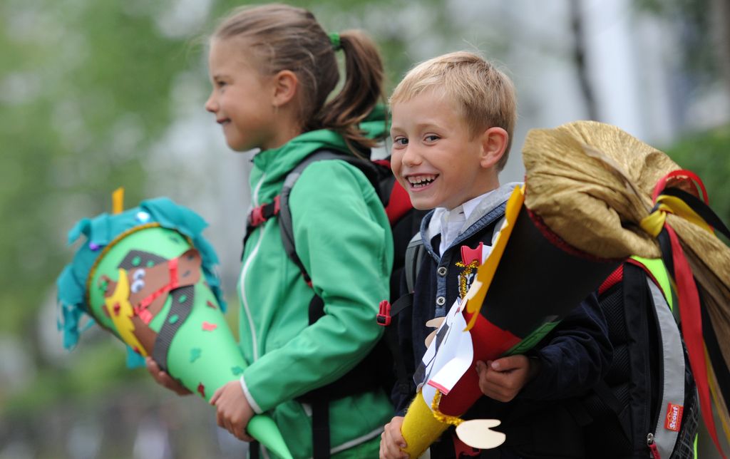 To smilande førsteklassingar, ein gut og ei jente, på veg til skulen første skuledag. Begge har ransel på ryggen og ber på enorme kremmerhus pynta med teikningar og pålimde motiv i mange fargar. Foto.