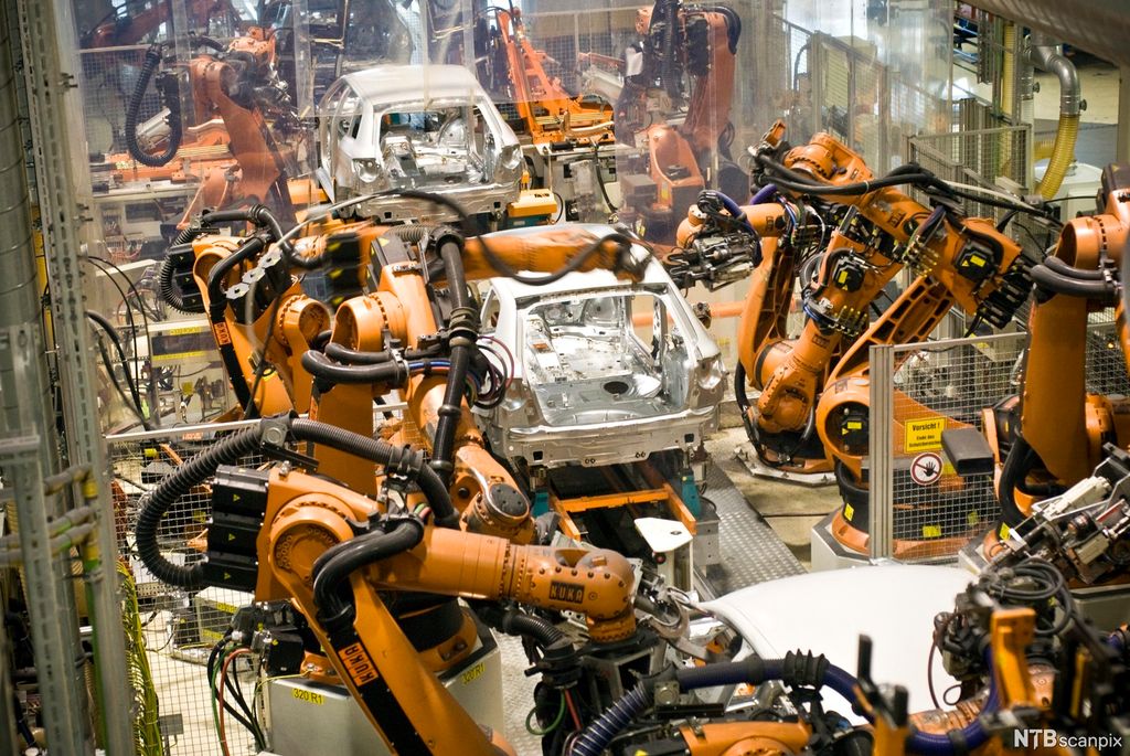 Produksjonslinje i en bilfabrikk. Tre ulakkerte, delvis sammensatte personbiler på et samleband. Til venstre og høyre for samlebandet kraftige oransjefarga robotarmer på rekke og rad. Foto.