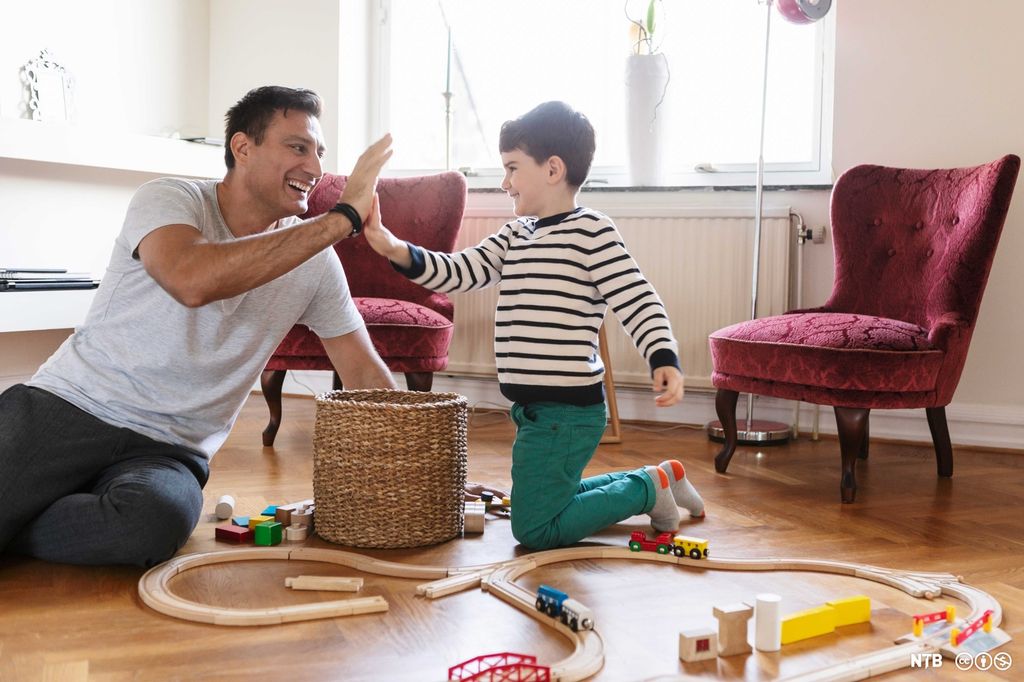 En mann og en gutt sitter på gulvet med en leketogbane. De smiler og gir hverandre high five. Foto.