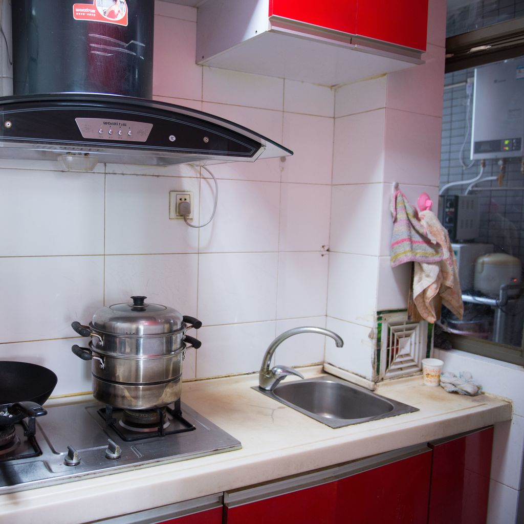 Noe slitt kjøkken med gassovn, kjøkkenvifte og en liten vask. Det er hvite fliser på veggene og røde fronter på innredningen. Foto.