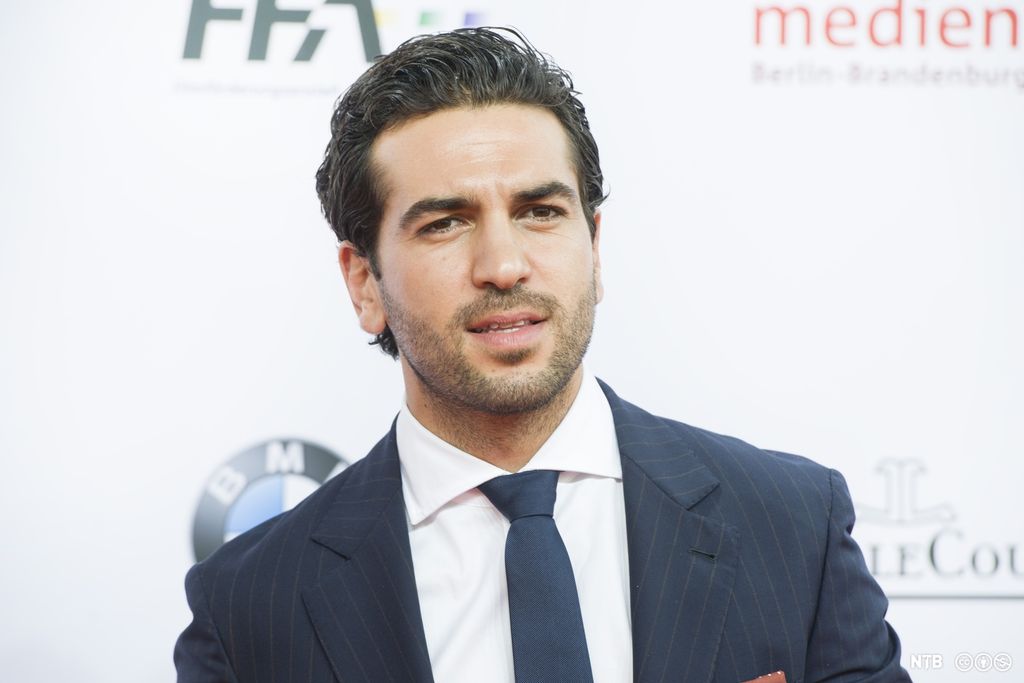 Nærbilde av en mann på rundt 30 år. Han har mørkt, kort hår, kort skjegg og brune øyne. Han har på seg mørkeblå dress, hvit skjorte og mørkeblått slips. Foto.