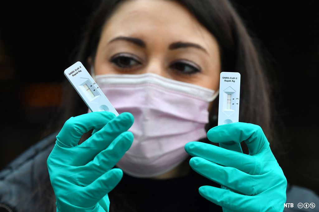 Kvinne med munnbind og grønne hansker holder i to hurtigtester for covid-19. Foto.