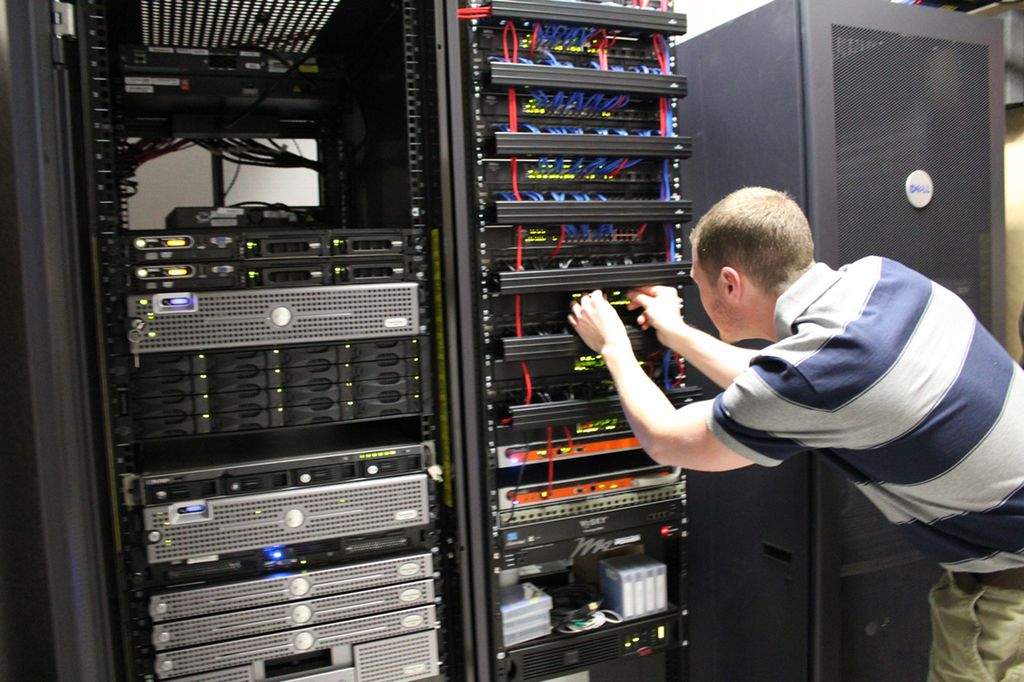 Flere høye metallskap med perforerte dører foran og bak. I metallskapene er servere stablet oppover. En mann står bøyd og kobler kabler til en svitsj i et av skapene. Foto.