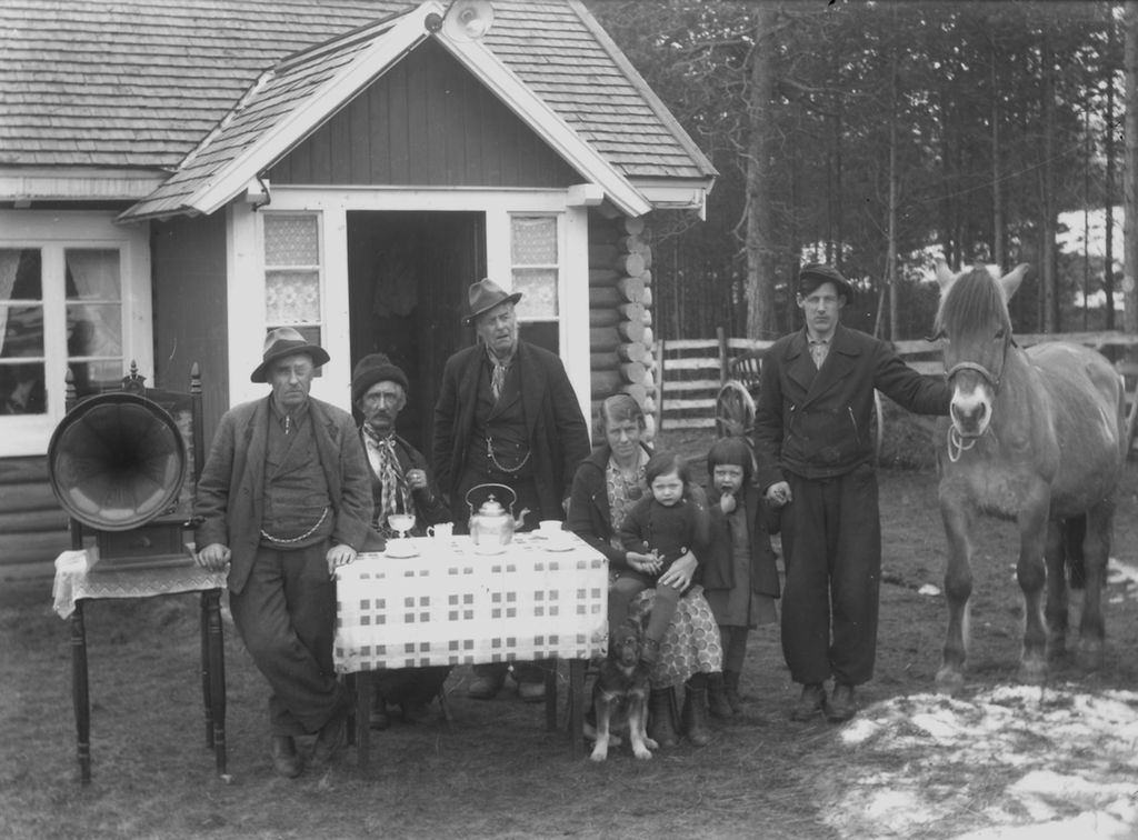 En familie med flere generasjoner sitter ved et kaffebord ute på tunet foran et hus. Den ene mannen holder en hest. En sveivegrammofon står ved siden av bordet. En hund er også med på bildet. Bak huset er det skog. Foto. 