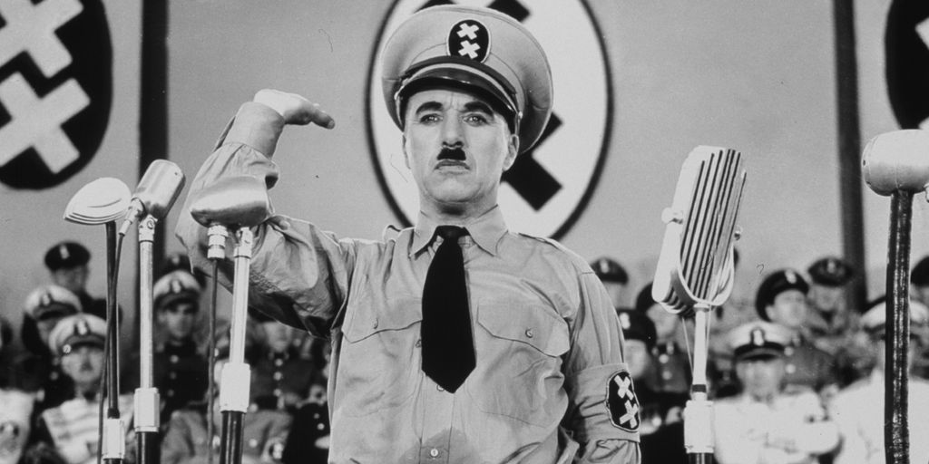 Chaplin med uniformslue og Hitler-bart gjør en forvridd nazihilsen. På lua og på veggen bak er det hakekorslignende symboler. Foran ham er det mange mikrofoner. Bak ham er det mange folk med lignende luer. Foto.