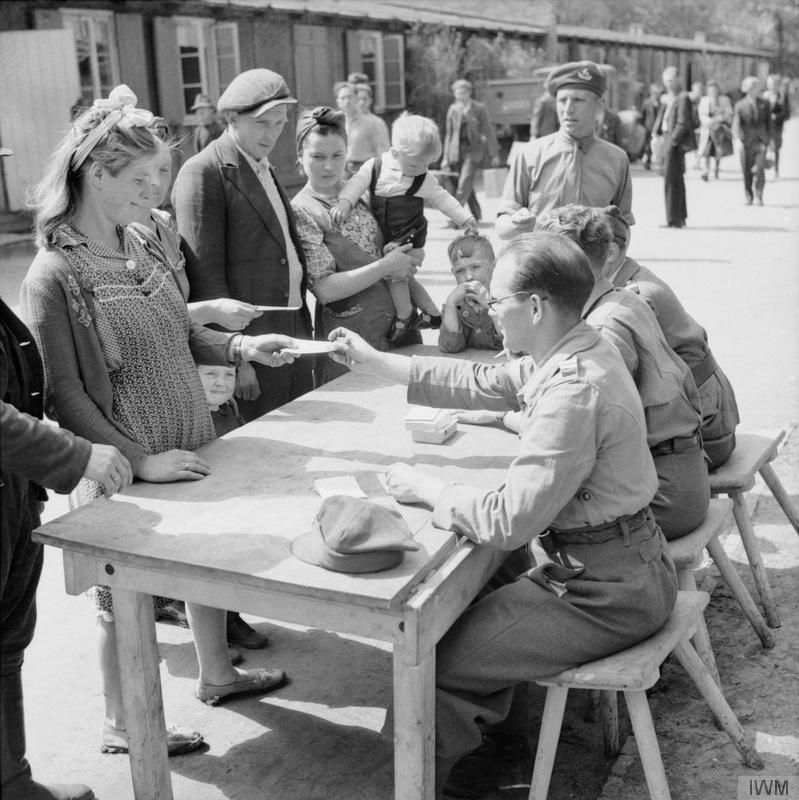 En familie står foran et bord på en plass ute foran en lang, brakkelignende bygning. Bak bordet sitter tre unge karer som ordner med papirer til familien. Foto. 
