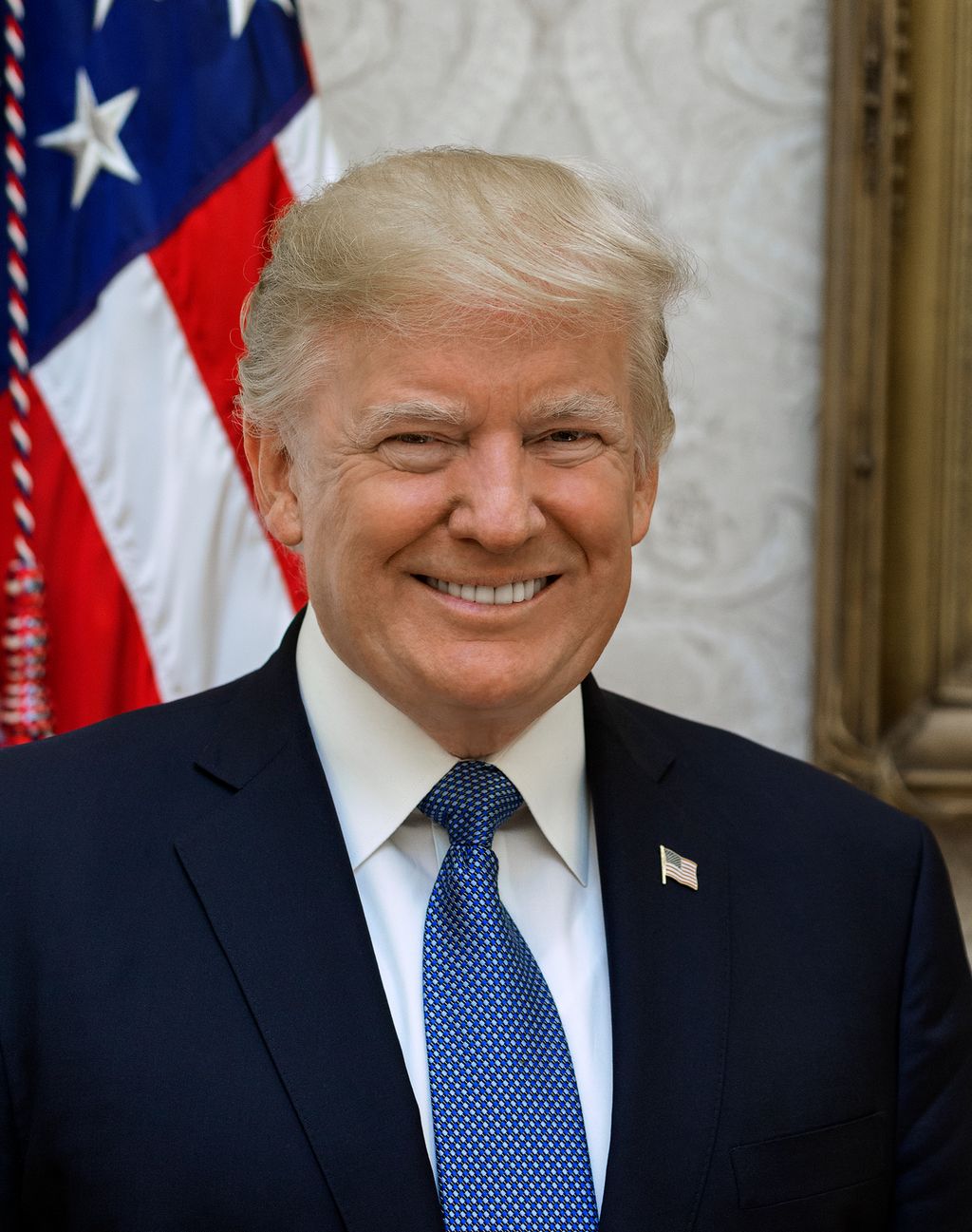 Portrait of President Donald Trump. He is wearing a blue suit, and a blue tie. He wears a flag pin. There is a flag behind him. Photo.